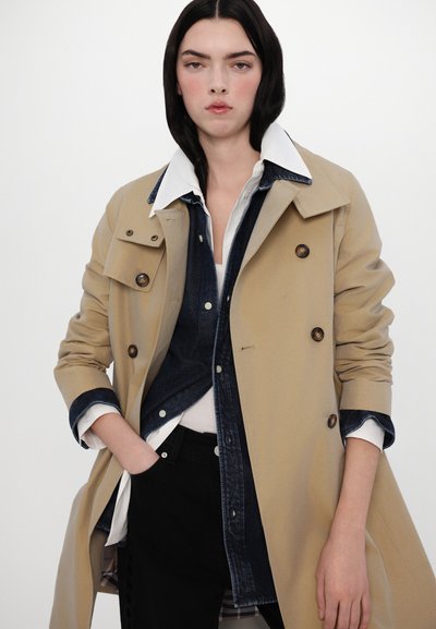 Cămașă trench bej cu design dublu, nasturi mari și guler alb contrastant peste o cămașă din denim bleumarin, asortată cu pantaloni negri.