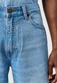 Jasnoniebieskie dżinsy mają wysoki stan, prosty krój i kontrastowe przeszycia. Z przodu znajdują się zapięcie na guzik i boczne kieszenie.