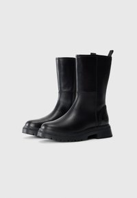 Et par svarte lærskaftboots til midten av leggen med tykk gummisåler og trekkremmer, plassert side om side på en enkel lys bakgrunn.