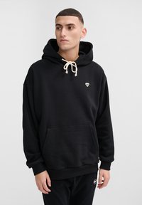 Svart hoodie med känguruficka, justerbara krämfärgade dragsnoddar och en liten vit logotyp på bröstet. Mjuk bomullsmaterial, avslappnad passform.