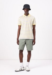 Polo t-shirt i gul og lyse lilla striber, parret med grønne shorts. Har en krave, broderet logo og grå sneakers.