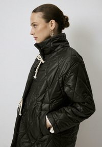 Chaqueta acolchada negra con cuello alto, que presenta costuras diagonales, detalles con cordón y puños blancos. Textura suave y ajuste relajado.