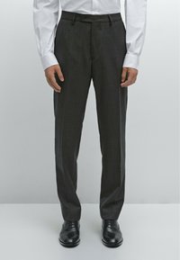 Dunkelgraue maßgeschneiderte Hose mit geradem Schnitt, mit einem Knopfverschluss und Taschen. Getragen mit einem weißen Langarmhemd und schwarzen Schuhen.