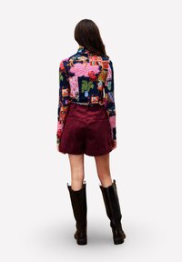 Vrouw met lang bruin haar, gekleed in een kleurrijke, geprinte top met lange mouwen, bordeaux rode shorts en zwarte kniehoge laarzen, staand met de rug naar de kijker.