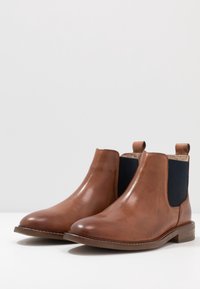 Bottines en cuir marron avec un bout rond, des panneaux latéraux élastiques bleu marine et un petit talon empilé marron. Présentent des détails de couture sur la partie supérieure.