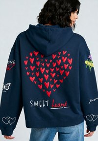 Navy hoodie met een groot rood hartontwerp gemaakt van kleinere harten, met geborduurde tekst "SWEET heart" en kleurrijke accenten op de mouwen.