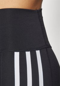 Leggings neri a vita alta con una texture liscia, caratterizzati da tre strisce bianche verticali lungo i lati e dettagli con cuciture flatlock.