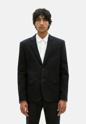 Schwarze Sakko mit zwei Knöpfen, aufgesetzten Taschen und Revers. Getragen über einem weißen Hemd mit Kragen, das eine glatte Textur aufweist.