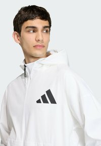 Biela kapucňov�á bunda z ľahkého materiálu, s čiernym logom Adidas na ľavej hrudi a predným zipsom.