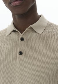 Beige polo póló, texturált hullám mintával, klasszikus gallérral és két fekete gombbal a nyakrészen.