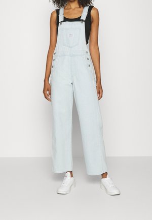Femme portant une salopette en jean bleu clair, un débardeur noir et des baskets blanches, debout les mains détendues le long du corps.