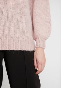 Jasnoróżowy sweter wykonany na drutach o luźnym kroju, z prążkowanymi mankietami i dołem, noszony na czarnych spodniach. Tekstura jest miękka z delikatnie puszystym wyglądem.