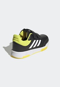 Zapatilla deportiva negra con rayas blancas y un cuello de malla amarillo. Cuenta con una correa de velcro y una suela de goma con acentos amarillos.
