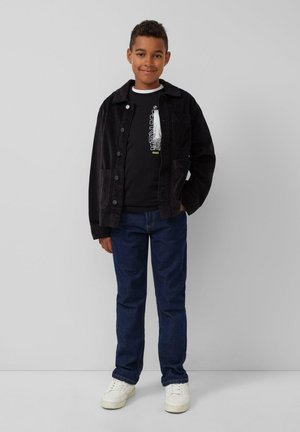 Zwarte corduroy jas met knopen aan de voorkant, gedragen over een zwart shirt met witte en gele grafische tekst, in combinatie met blauwe spijkerbroeken en witte sneakers.