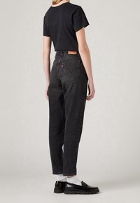 Personne portant un jean noir taille haute, un t-shirt noir, des chaussettes blanches et des mocassins noirs, debout devant un fond clair uni.