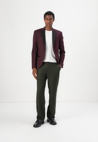 Bordeauxfarbener Blazer mit schwarzen Revers, weißem Hemd, grüner Hose und schwarzen Schuhen. Schmal geschnitten mit glatter Textur und maßgeschneidertem Design.