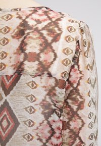 Blusa transparente con un patrón geométrico multicolor en tonos de marrón, rosa y crema. Detallada con pliegues en los hombros.