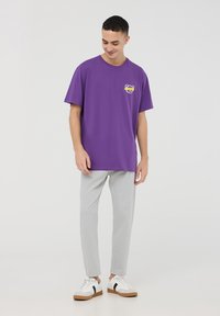 T-shirt di cotone viola con collo rotondo, caratterizzata da una grafica gialla sul petto. Abbinata a pantaloni aderenti grigio chiaro e sneakers bianche.