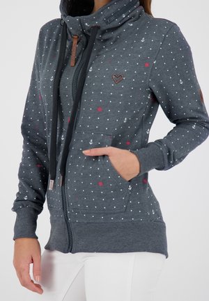 Sweat à capuche zippé gris foncé avec motif de pois blancs et rouges et ancres, poche avant et cordons noirs, porté avec un pantalon blanc.