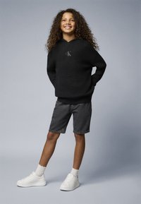 Czarna dziana bluza z kapturem z haftowanym logo "ck", połączona z szarymi szortami dżinsowymi i białymi sneakersami, uzupełniona strukturalnymi białymi skarpetkami.