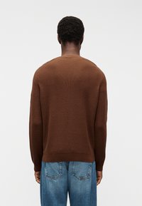 Moški s kratkimi kodrastimi lasmi, oblečen v rjav dolg rokav pulover in modre jeans hlače, stoji obražen stran, na belem ozadju.