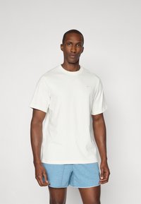 Camiseta blanca de manga corta con cuello redondo y un pequeño detalle de logo en el pecho, combinada con pantalones cortos de color azul claro.
