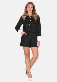 Conjunto de traje de tweed negro con chaqueta que presenta hilos metálicos, botones redondos y puños blancos, combinado con pantalones cortos a juego y tacones blancos.