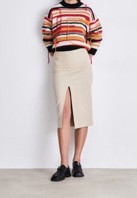 Pull en maille rayé rouge, orange et noir associé à une jupe beige longueur genoux avec une fente à l'avant et des chaussures noires à boucles.