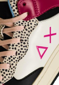 Sneakers con una base in pelle bianca, dettagli rosa, tessuto nero e sezioni in suede beige maculate. Presentano grafiche rosa e nere e lacci rosa.