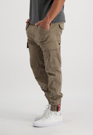 Mand iført beige cargobukser og hvide sneakers, hænder i lommerne, står mod en lys ensfarvet baggrund.
