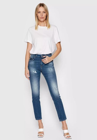 Donna bionda che indossa una semplice maglietta bianca, jeans blu strappati e tacchi aperti bianchi, in piedi contro uno sfondo chiaro.