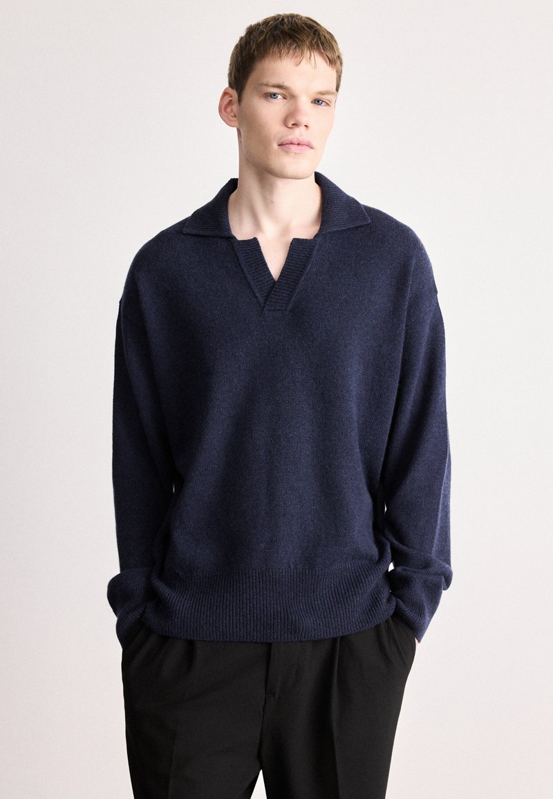 Πλεκτό πουλόβερ σε ναυτικό μπλε με V-λαιμόκοψη και φαρδιά γιακά. Μαλακή υφή και ριμπ μανσέτες. Φορεμένο πάνω από μαύρο παντελόνι.