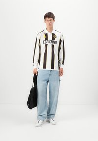 Langermet, hvit fotballdrakt med svarte vertikale striper og gule detaljer, med teksten "F.C.TECHNO". Brukt med lyseblå baggy jeans.