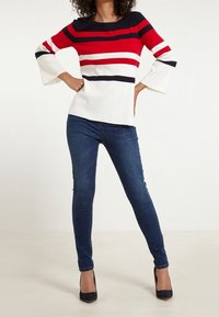 Langarmtop mit roten, weißen und marineblauen Streifen und ausgestellten Ärmeln, kombiniert mit dunkelblauen Skinny Jeans und schwarzen High Heels.