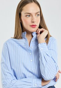 Chemise boutonnée à rayures bleu clair avec un col haut, une patte de boutonnage, et des manches longues présentant des poignets boutonnés et des plis subtils.