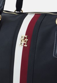 Sac en tissu marine avec des rayures blanches et bordeaux, matériel doré et accent de logo. Texture lisse avec détail de fermeture éclair.