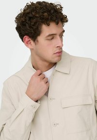 Veste beige boutonnée avec col, poche avant et manches retroussées, en tissu lisse. Le modèle tient délicatement le col.