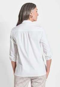 Weißes Hemd mit aufgerollten Ärmeln und gerüschtem Rückenpasse. Kombiniert mit hell gemusterten Hosen und goldenen Ohrringen.