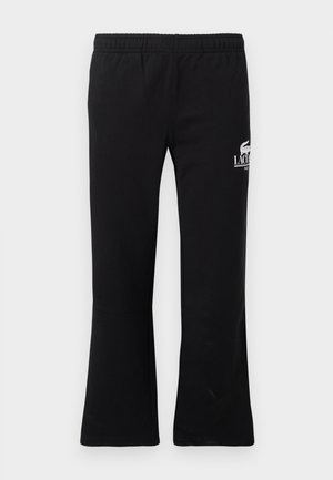 Deux adultes portant des chemises blanches à manches longues et des pantalons de survêtement noirs Lacoste se tiennent contre un mur blanc, chaussés de baskets blanches.