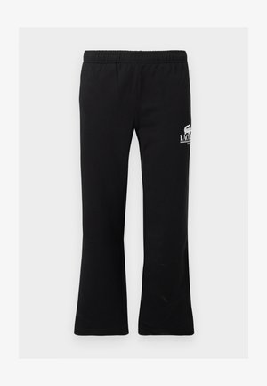 Deux adultes portant des chemises blanches à manches longues et des pantalons de survêtement noirs Lacoste se tiennent contre un mur blanc, chaussés de baskets blanches.