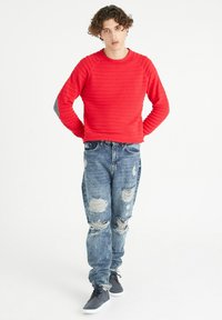 Suéter rojo texturizado con diseño acanalado, combinado con jeans azul desgastados que presentan desgarros. El modelo lleva zapatillas grises.