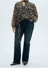 Blouse fleurie noire et crème avec un lien au col, associée à un jean flare bleu foncé avec des fentes sur les côtés. Chaussures noires à bouts pointus.