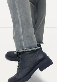 Botas de cuero navy hasta el tobillo con acabado texturizado, cierre de cordones y suela robusta, combinadas con unos pantalones vaqueros de denim azul claro con dobladillos deshilachados.