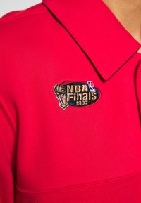 Polo rouge avec col, arborant un logo brodé des Finales NBA 1997 en or, noir et bleu. Texture de tissu lisse.