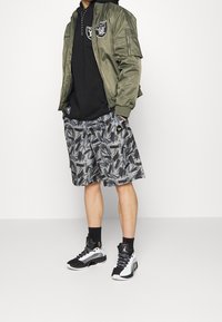 Olivgrön bomberjacka med svart hoodie under, grå mönstrade shorts och svarta och vita sneakers. Funktioner inkluderar logotyper och textdetaljer.