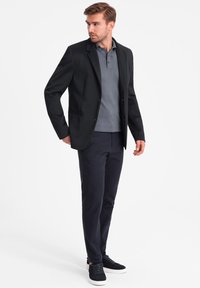 Schwarzer Blazer über einem grauen Poloshirt, kombiniert mit navyfarbener Hose und schwarzen Sneakers. Glattes Material, maßgeschneidertes Design und schlanke Silhouette.