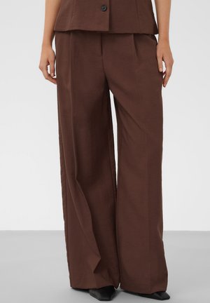 Femme portant un pantalon marron à jambes larges et un gilet marron assorti boutonné, debout devant un fond clair uni.