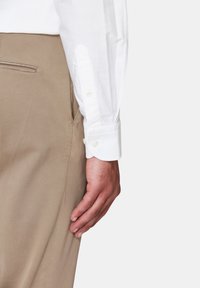 Camicia bianca a maniche lunghe con polsini bottonati, abbinata a pantaloni beige chiaro dotati di tasca laterale e una texture di tessuto liscia.