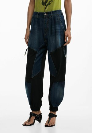 Kobieta ubrana w luźne, patchworkowe dżinsy oraz czarne spodnie dresowe z zamkami, elastycznymi ściągaczami i czarne sandały na obcasie.