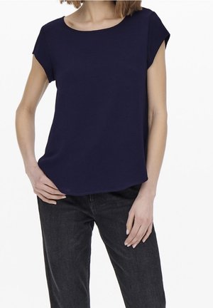 Kvinde iført en mørkeblå kortærmet top og sorte jeans, stående med den ene hånd på hoften og den anden hviler ved siden.